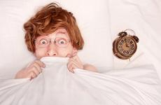 A redheaded man in glasses is laying in bed and pulls a white sheet up to cover his mouth in surprise while an alarm clock rests on the pillow beside him.