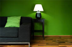 A black couch with a green pillow sits next to a black end table with a black lamp and white shade in front of a green wall. 