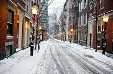 A snowy city street lined on either side with brick multistory buildings and street lights.