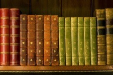 Rows of old-looking hardback bound books in red, brown, orange, and green across a wooden bookshelf. 