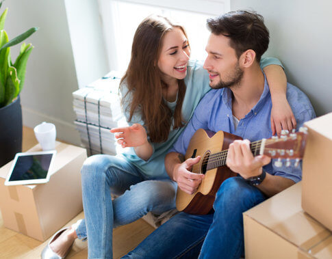 Young smiling couple surround by moving boxes, playing guitar and making memories in their new apartment