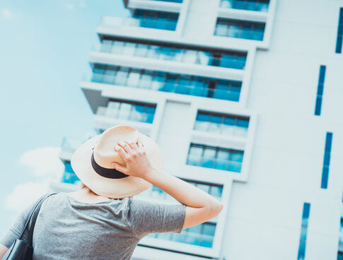 Person with fedora hat is apartment hunting and staring up at tall white apartment complex building