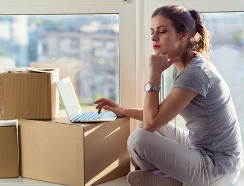 Woman sitting on floor of her apartment with laptop on stack of cardboard boxes, as she considers moving to a new apartment