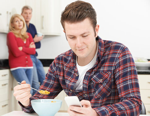 Adult son eating breakfast at his parent's house, with parents looking frustrated in the background