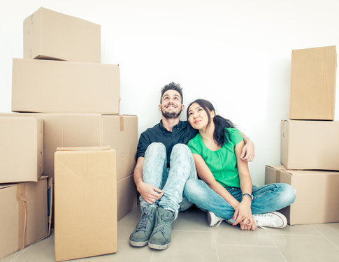 Young couple moving into new apartment, sitting on floor surrounded by boxes but looking excited