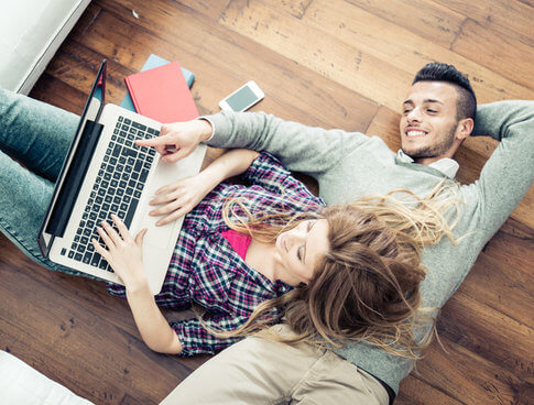 Happy couple laying on floor, looking at apartment search sites on laptop
