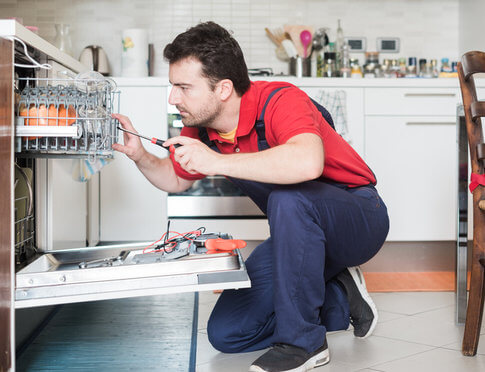 Apartment repairman in blue overalls and red t-shirt with screwdriver, fixing apartment dishwasher