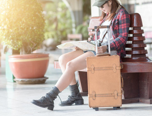 Young woman in ball cap sitting on a bench with trendy suitcase, looking at a map