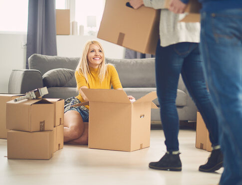 Blonde girl in yellow shirt unpacking moving boxes, with other roommates arriving