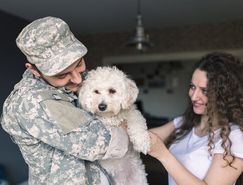 Military man holding fluffy white dog, next to wife looking on with joy