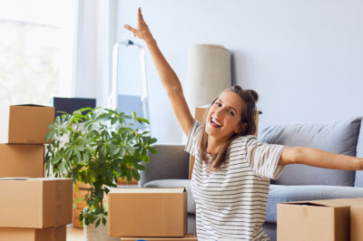 A young woman is happy with outstretched arms surrounded by moving boxes in new apartment.