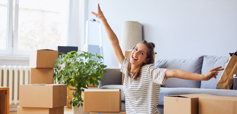 A young woman is happy with outstretched arms surrounded by moving boxes in new apartment.