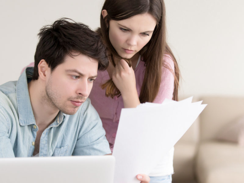 A young couple on a laptop sits on a light colored couch looking at paperwork.