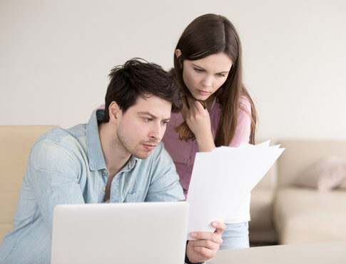 Couple in front of computer in apartment, looking confusedly at papers
