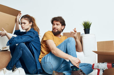 Young couple sits on couch with moving boxes facing away from one another in visible argument.