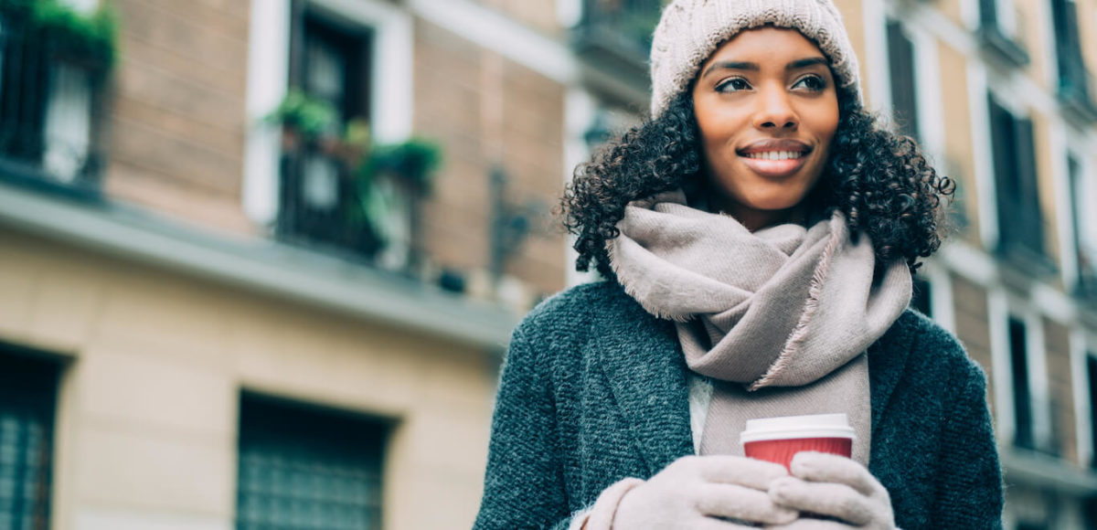 Young woman on street wearing winter hat, gloves, and scarf holds coffee while smiling.