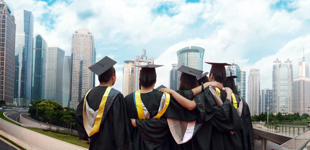 A group of graduates in cap and gown stand with their backs to the camera overlooking the city.