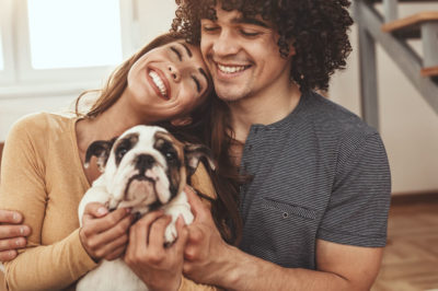 A young couple nestles together with moving boxes in the background holding a bulldog puppy.
