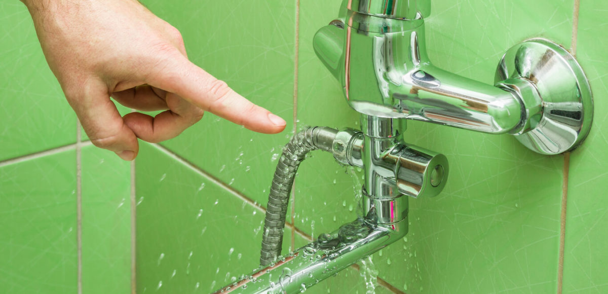 A mans hand points to a leaking shower hose in a green tiled shower.