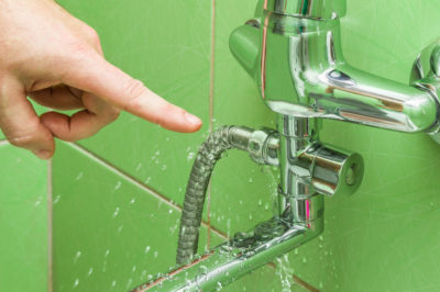 A mans hand points to a leaking shower hose in a green tiled shower.