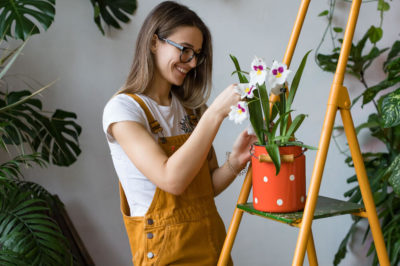 A woman in overalls is surrounded by plants and tends to an orchid in an orange pot on a step ladder.