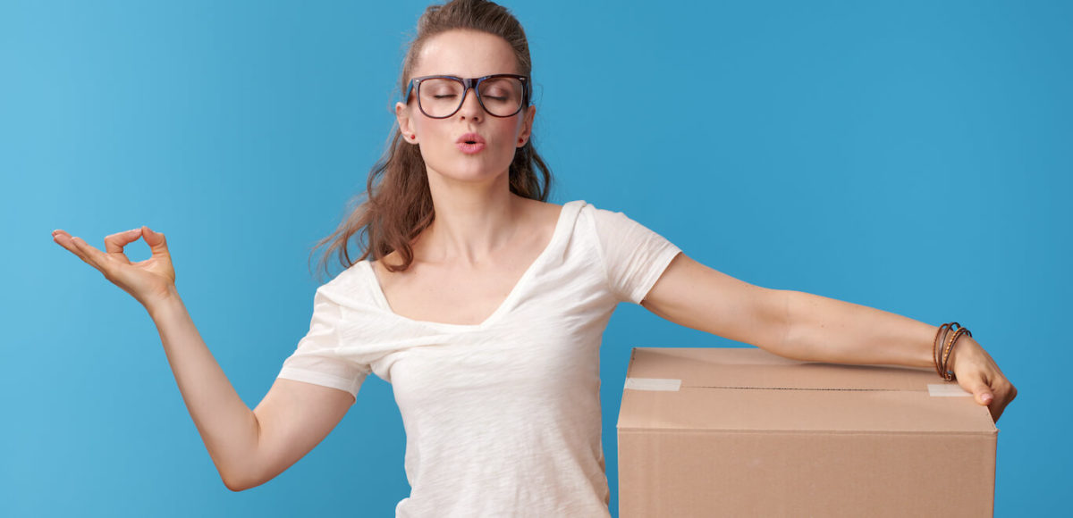 A woman in a white T shirt and red flannel tied around her waist makes the om symbol with one hand while cradling a moving box to her hip.
