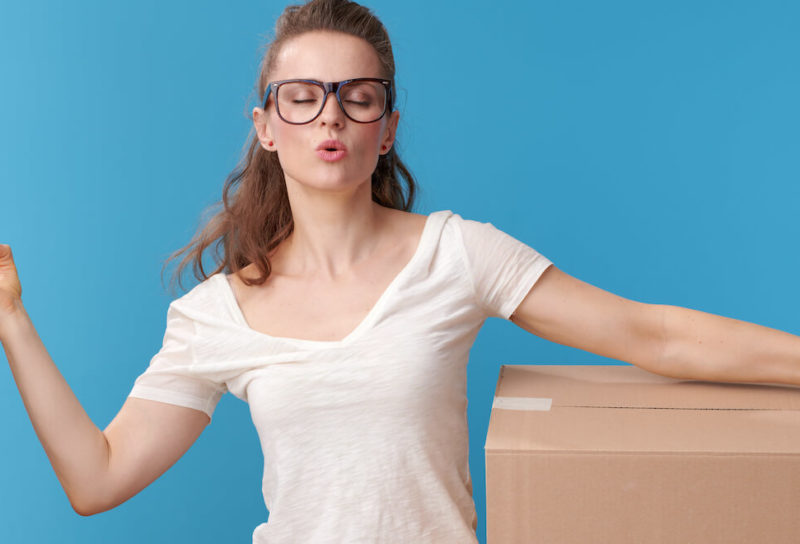 A woman in a white T shirt and red flannel tied around her waist makes the om symbol with one hand while cradling a moving box to her hip.