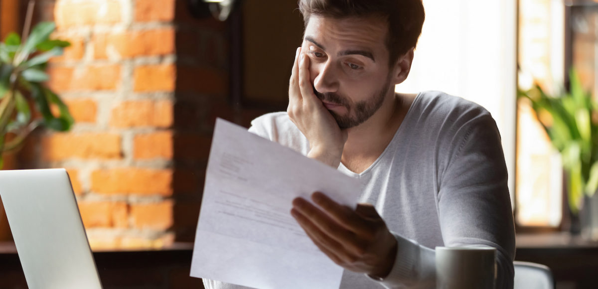 A man in a brick walled apartment sits staring disappointedly at a document.