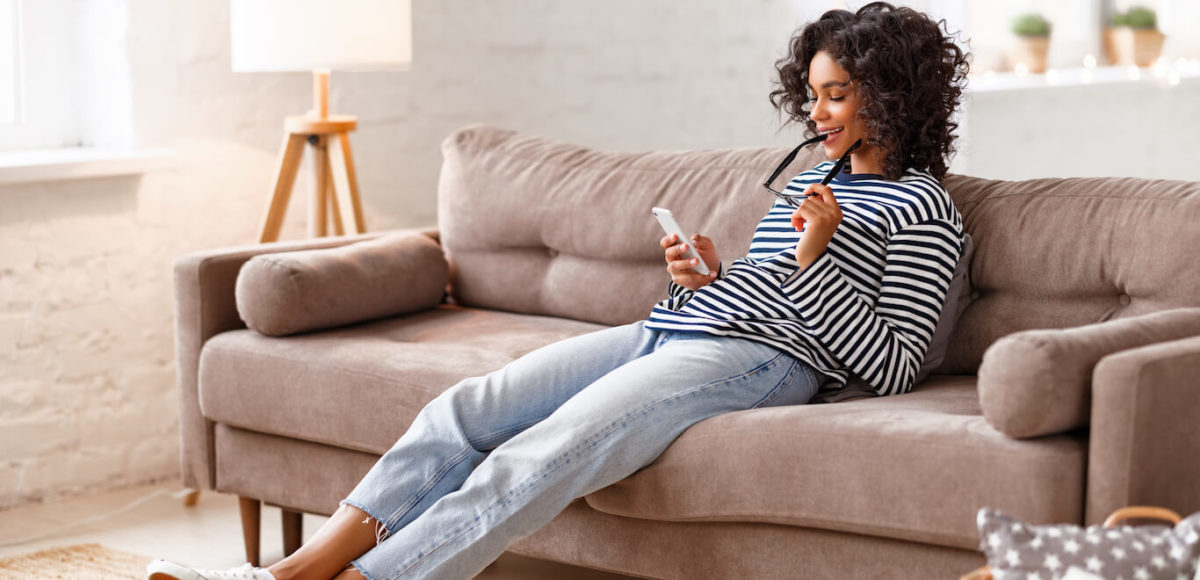 A woman sits on the couch with legs outstretched smiling as she checks her phone.