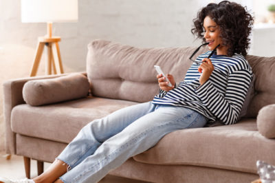 A woman sits on the couch with legs outstretched smiling as she checks her phone.