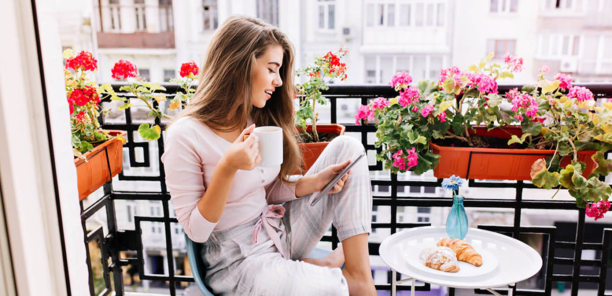 Woman in pajamas sits on balcony at small table checking her phone surrounded by pink potted flowers.