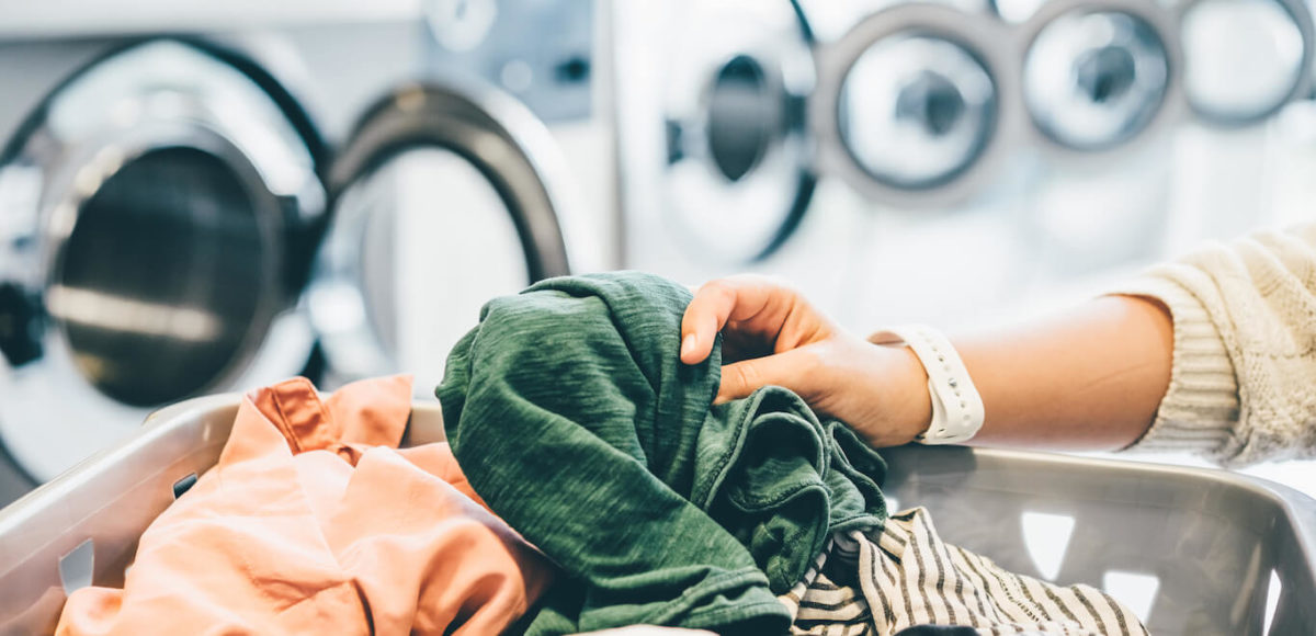 A woman's hands grab dirty laundry from a grey laundry basket to place in the washing machine.