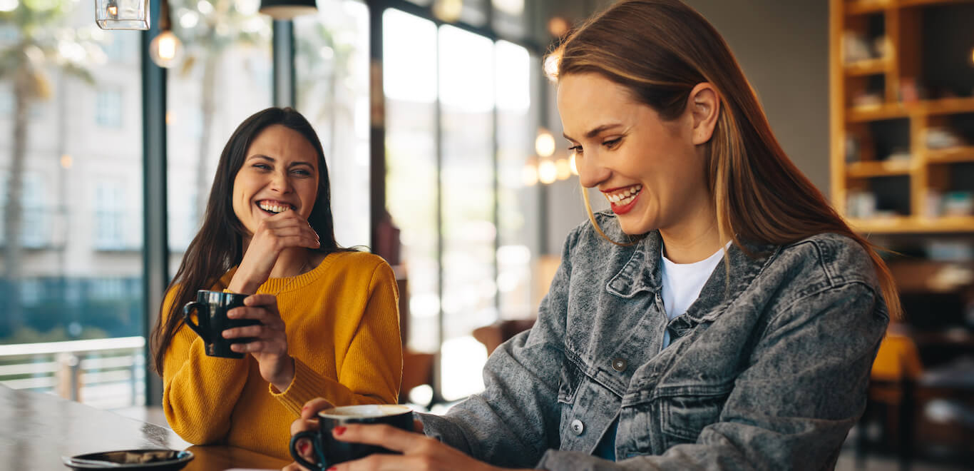 Two young women sit at countertop drinking from black coffee mugs and smiling.