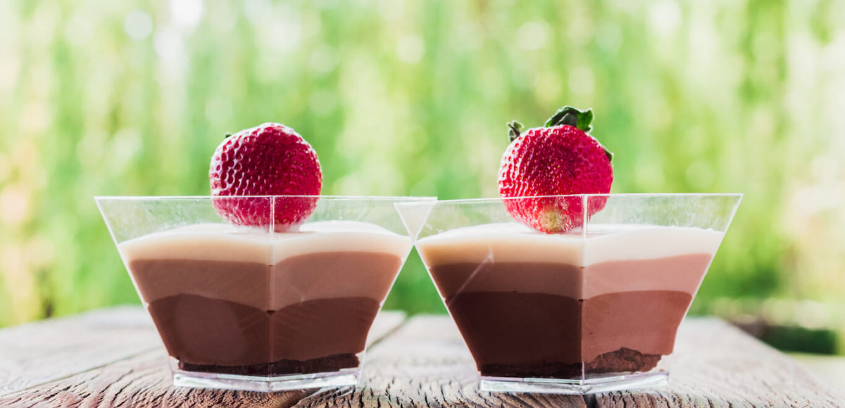 Two chocolate pudding parfaits topped with a strawberry each sit on a wood planked surface with greenery in the background.