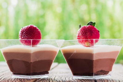 Two chocolate pudding parfaits topped with a strawberry each sit on a wood planked surface with greenery in the background.