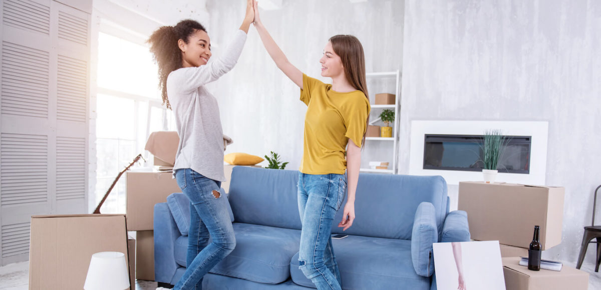 Two young women high five in living room in front of blue couch with moving boxes surrounding them.