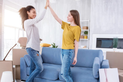 Two young women high five in living room in front of blue couch with moving boxes surrounding them.