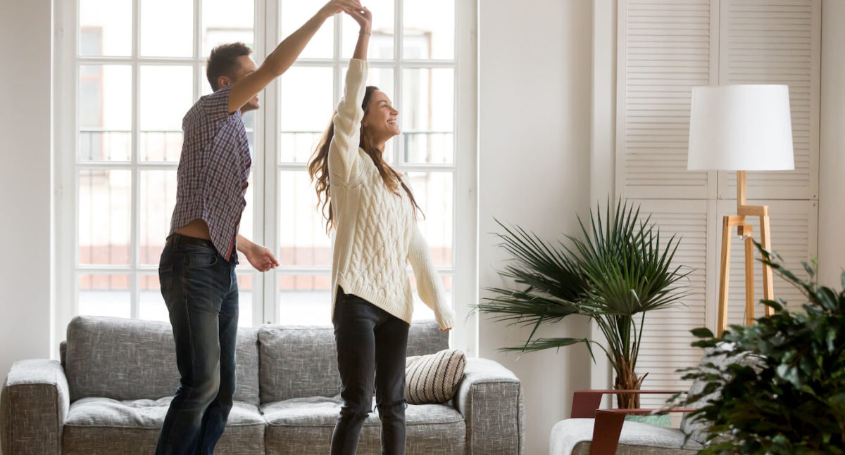 Young couple stands in living room in front of grey couch as he twirls her.