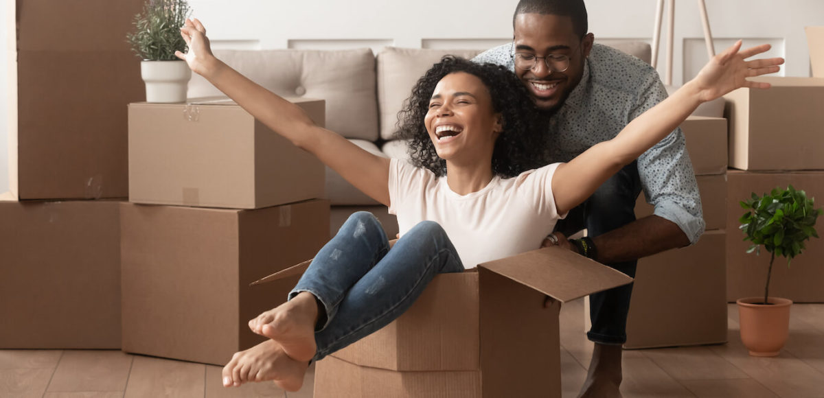 Two first time home buyers are having fun unpacking and laughing on moving day, excited woman is sitting in cardboard box being pushed by a man.