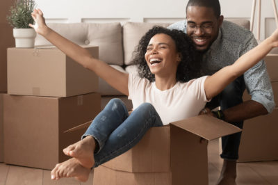 Two first time home buyers are having fun unpacking and laughing on moving day, excited woman is sitting in cardboard box being pushed by a man.