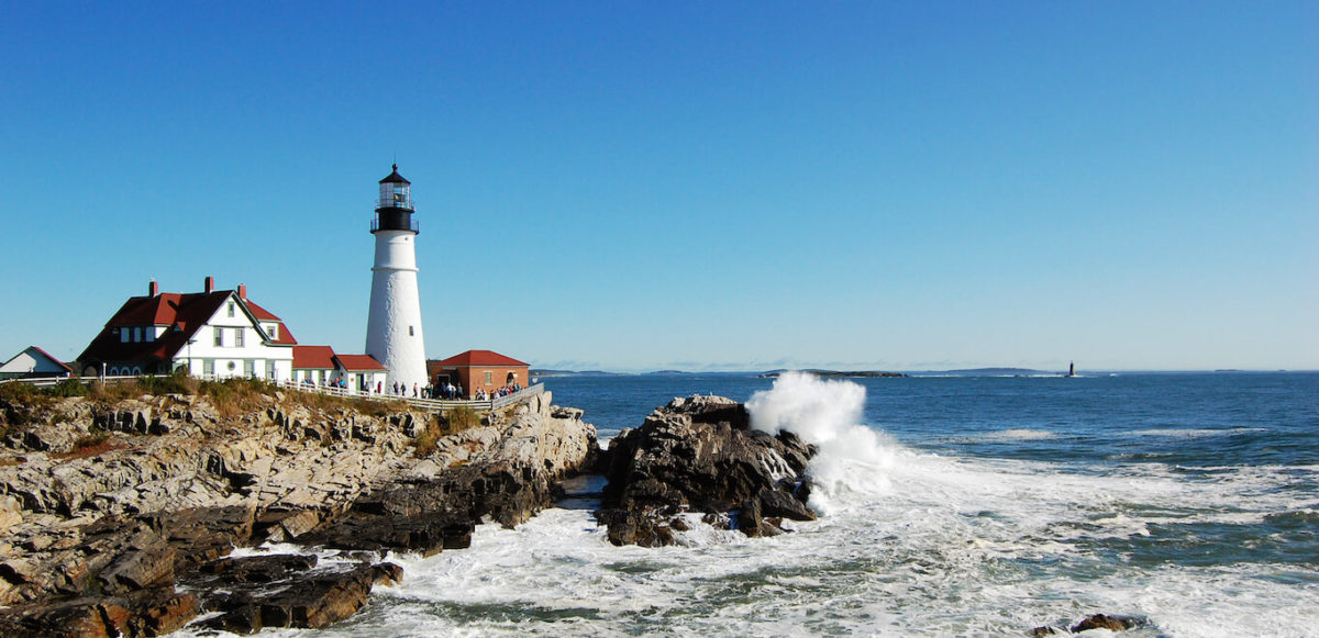 Landscape shot of Portland Head Lighthouse in Maine with big cloudless blue sky, white lighthouse, and waves breaking on the rocks.