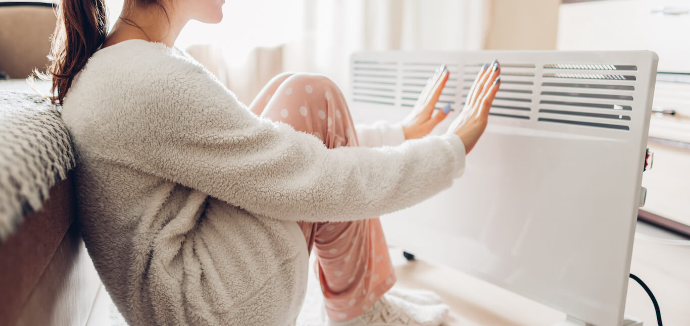 Woman in comfortable pajamas sits in front of white portable space heater with her hands extended.