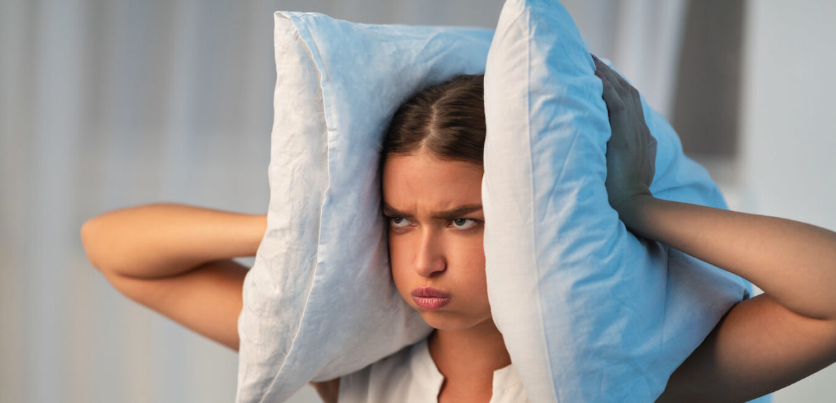 Young woman in pajamas presses two pillows to her ears in apparent frustration.