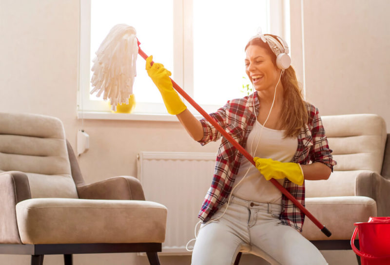 Young woman on her knees in living room with modern furnishings plays air guitar with a mop while wearing yellow cleaning gloves and white headphones.