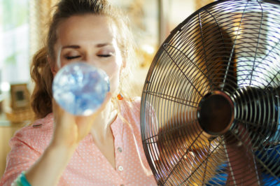 Young woman in pink button up top wearing ponytail drinks from a plastic water bottle in front of a metal fan in an attempt to cool down.