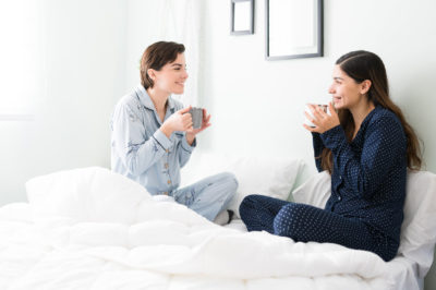 Two women in pajamas sit on a bed drinking coffee while having a pleasant conversation.