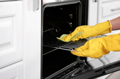 Woman in yellow rubber gloves scrubs stainless steel oven in kitchen with white cabinets.