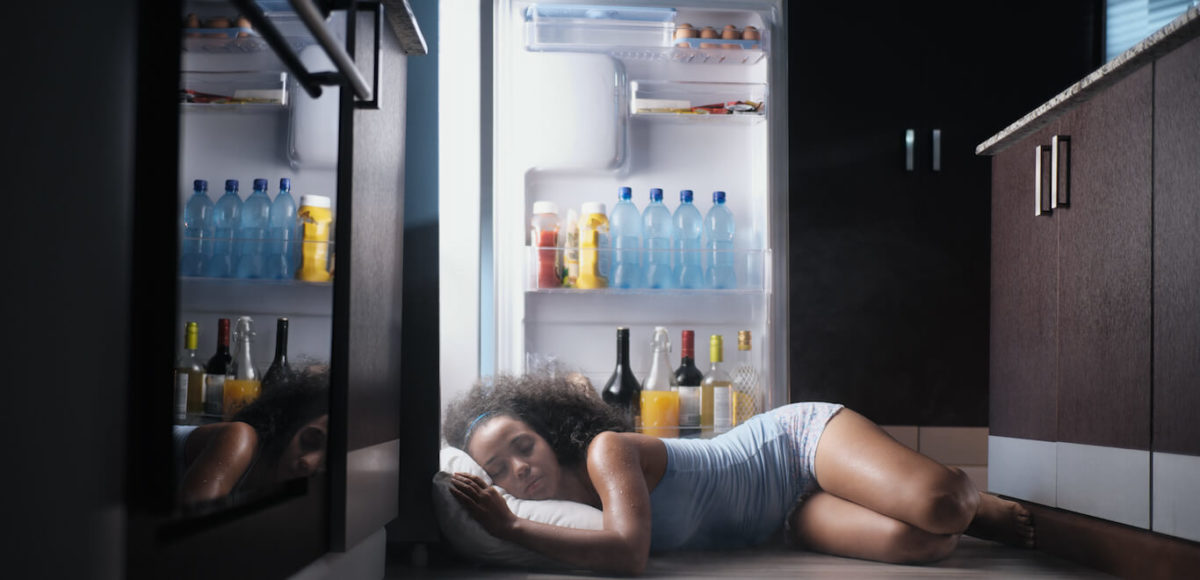 Woman sleeps on hardwood floor in kitchen with her head nearly in the open refrigerator in an attempt to cool down.