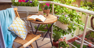 An inviting balcony featuring a wood slatted chair with a blanket and pillow, a table with an open book, and an assortment of greenery,