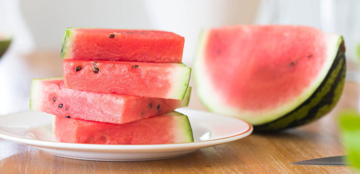 Watermelon slices rest on a white plate while a quarter of a watermelon sits aside it on a maple colored countertop.